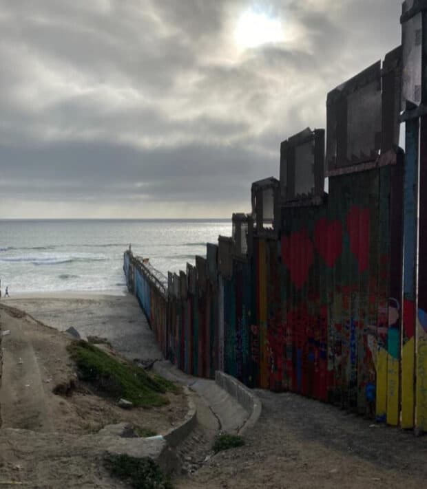 Painted border fence extending into the ocean under a cloudy sky.
