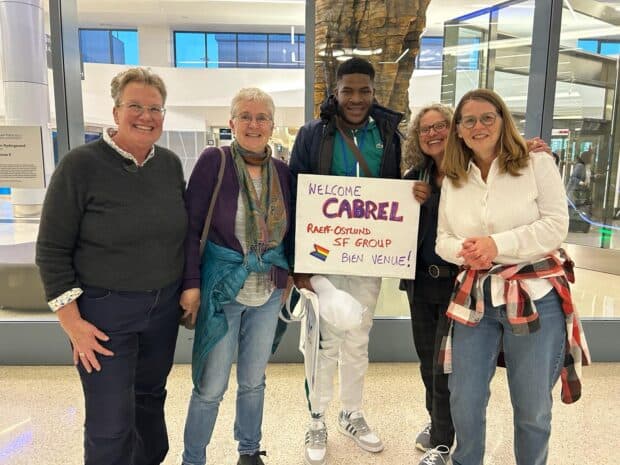 A group of five people at an airport holding a welcome sign and smiling together.