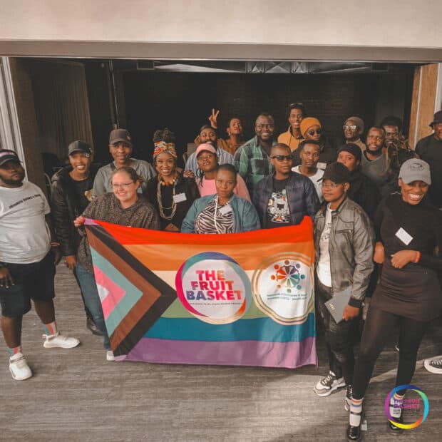 Group photo indoors holding a Pride flag that reads “THE FRUIT BASKET.”
