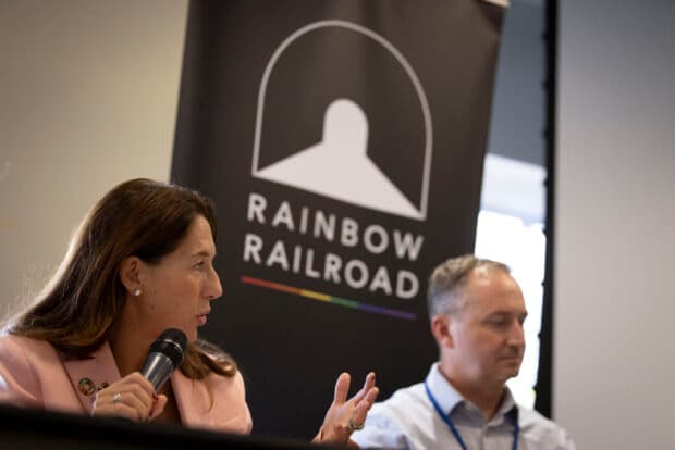 Two panelists speak beside a Rainbow Railroad banner at a formal event.