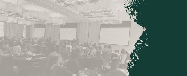 People seated at round tables in a conference room listening to a presentation on large screens.