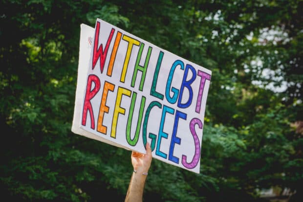 Hand holding a rainbow-coloured sign reading “WITH LGBT REFUGEES.”