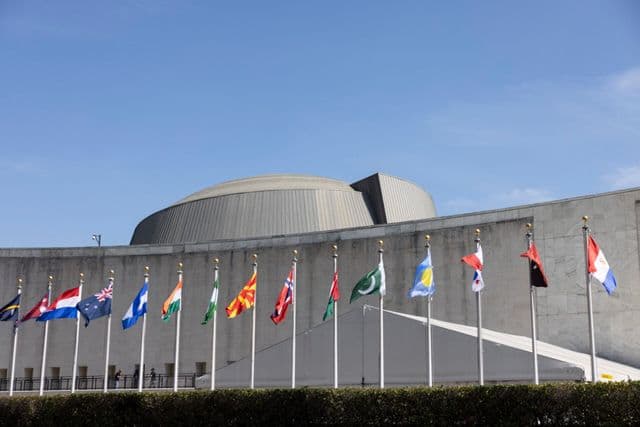 Row of international flags flying in front of a concrete building.
