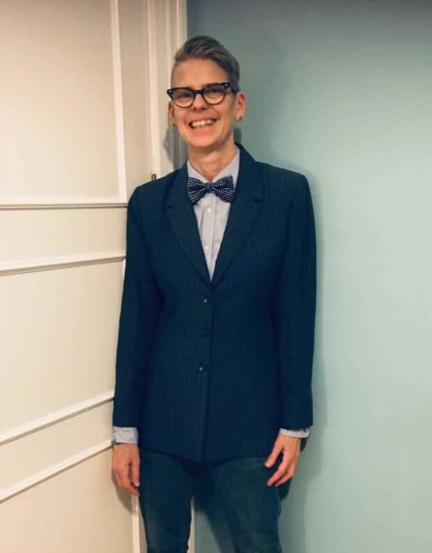 Person smiling while wearing a suit and bow tie, standing indoors.