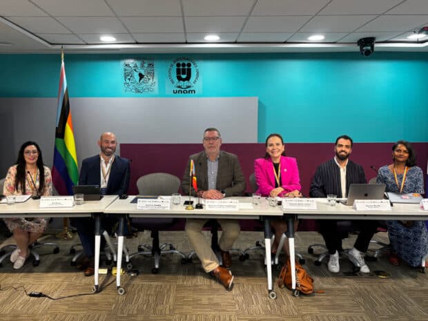 Six panelists seated at a long table with microphones and laptops, in front of a teal wall and a rainbow flag.