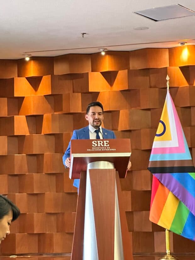 Person at a podium speaking beside a Pride flag in a wood-panelled room.