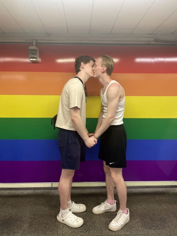 Couple kissing in front of a wall painted with rainbow stripes, holding hands and facing each other.