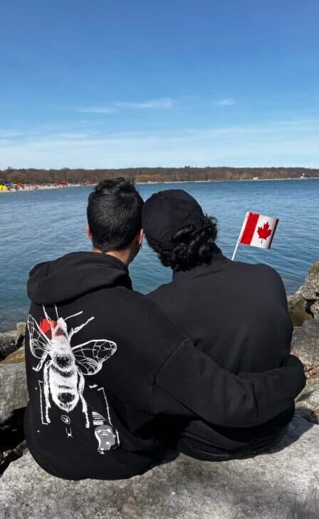 Two people sitting on a bench by the water, one resting their head on the other's shoulder, holding a Canadian flag.