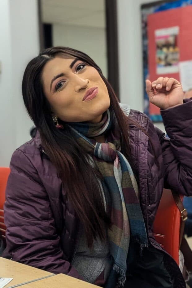 Person sitting indoors in a scarf and jacket, smiling.