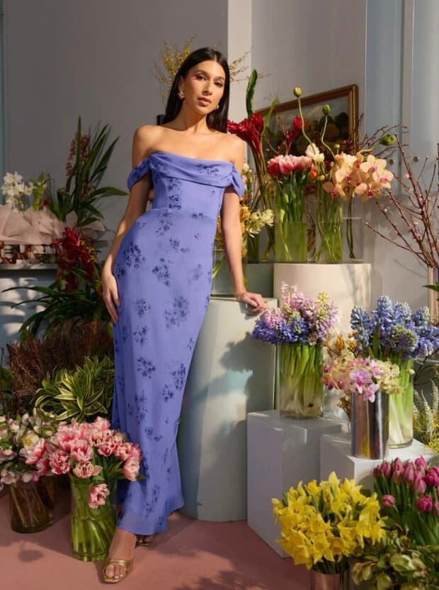 Person in a lavender off-shoulder floral dress standing among colorful flowers.