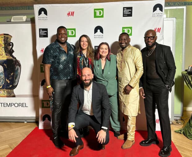 Group of six people smiling on a red carpet in front of a Rainbow Railroad Freedom Party backdrop.