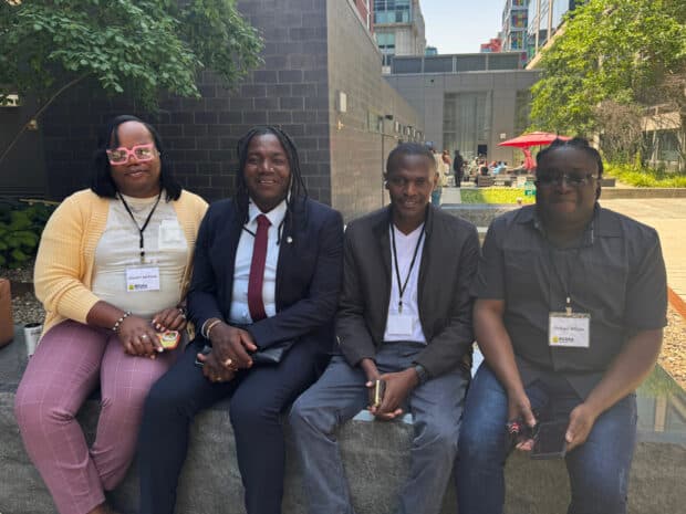 Four people seated outdoors on a ledge smiling, wearing conference badges.