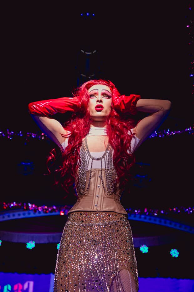 Drag performer with red hair and gloves posing onstage under bright lights.