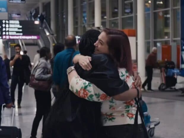 Two people hugging in an airport arrivals area, one wearing a floral top and smiling warmly.